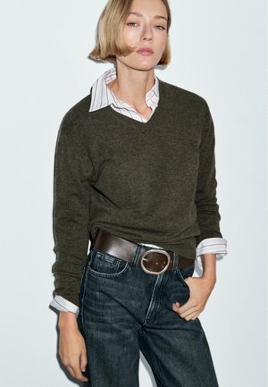 Dunkelgrüner Wollpullover mit V-Ausschnitt, über einem weißen Hemd mit pinken Streifen getragen. Kombiniert mit dunklen Denim-Jeans und einem braunen Leder-Gürtel.