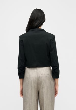 Une femme se tient de dos et présente un blazer noir court aux manches froncées et un pantalon beige à fines rayures, sur un fond uni.