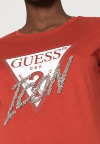 Primer plano de una camiseta roja con un gran logotipo de Guess en blanco y rojo y la palabra "ICON" escrita en cuentas plateadas en la parte frontal.