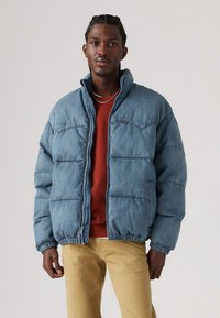 Denim pufferjakke i lys blå, med lynlås, h�øj krave og elastiske manchetter. Båret over en rød skjorte og khaki bukser.
