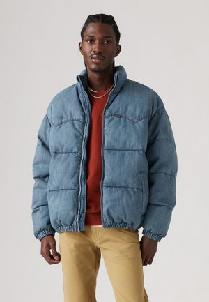 Denim puffer jacket in light blue, featuring a zip closure, high collar, and elastic cuffs. Worn over a red shirt and khaki pants.