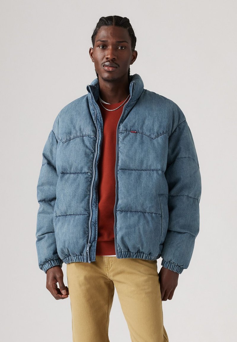 Denim pufferjakke i lys blå, med lynlås, høj krave og elastiske manchetter. Båret over en rød skjorte og khaki bukser.