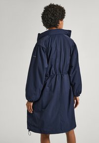Manteau long bleu marine avec taille à cordon, manches surdimensionnées et col montant. Fabriqué en tissu léger et texturé.