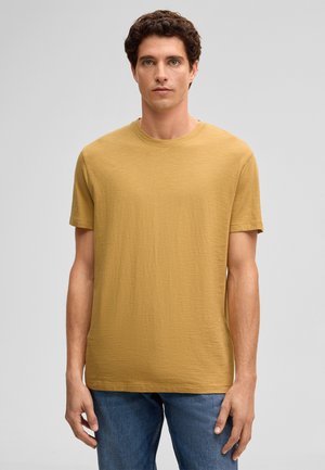 Jonge man met krullend donker haar, gekleed in een mosterdgele korte mouwen T-shirt en blauwe jeans, staande tegen een effen lichtgrijze achtergrond.