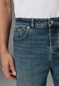 Blugi din denim albastru închis cu o ușoară decolorare, având un design clasic cu cinci buzunare, cusături în contrast și un sistem de închidere cu buton metalic.