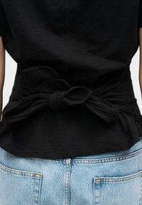 Μαύρο πουκάμισο με δεμένο υφασμάτινο φιόγκο στο κάτω μέρος της πλάτης, φορεμένο με ανοιχτό μπλε τζιν παντελόνι που δείχνει τις πίσω τσέπες και τις ραφές.