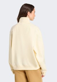 Sweatshirt crème avec col haut, coupe surdimensionnée et poignets côtelés. Texture lisse sans motifs, associé à un pantalon marron clair.
