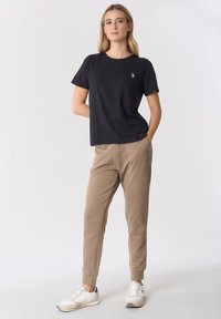 T-shirt nera in cotone con scollo rotondo e piccolo logo sul petto, abbinata a pantaloni della tuta beige e sneakers bianche con dettagli grigi.