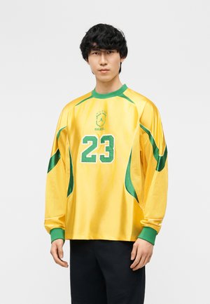 Νέος άνδρας με κίτρινη και πράσινη φανέλα της Βραζιλίας με τον αριθμό 23, στέκεται μπροστά σε λευκό φόντο.