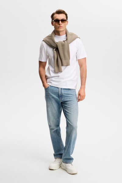 Άνδρας με γυαλιά ηλίου, λευκό μπλουζάκι, ανοιχτό μπλε τζιν, λευκά αθλητικά παπούτσια, με μπεζ πουλόβερ περασμένο στους ώμους, πάνω σε απλό φόντο.