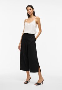 Camiseta sin mangas de encaje blanco combinada con pantalones anchos negros de cintura alta y aberturas laterales. La modelo lleva sandalias de tacón negro.