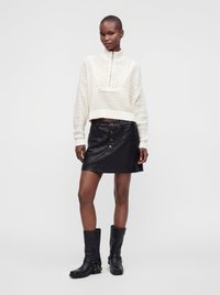 Femme portant un pull en maille blanc avec une fermeture éclair, une jupe en cuir noire boutonnée, et des bottes noires mi-mollet, debout sur fond blanc.