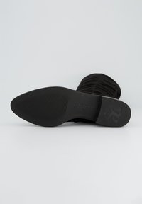 Botín negro de ante a nivel del tobillo con un diseño texturizado y acanalado. Suela de goma plana que muestra estrías verticales y un logo. Forma simple y elegante.