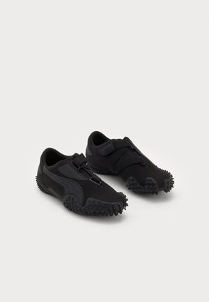 Paire de chaussures de sport noires avec semelles cloutées texturées et bandes Velcro sur fond blanc.