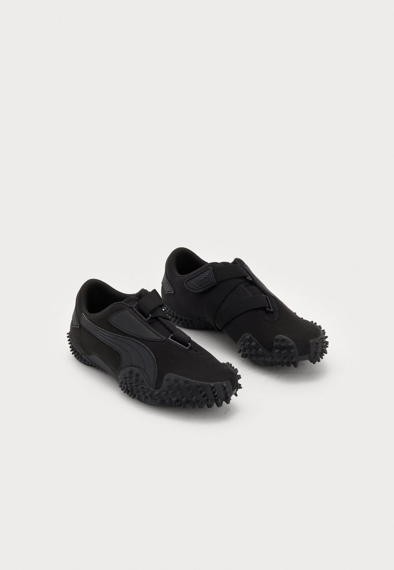 Paire de chaussures de sport noires avec semelles cloutées texturées et bandes Velcro sur fond blanc.