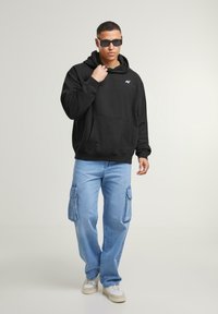 Sudadera con capucha negra y bolsillo frontal, combinada con pantalones cargo azul claro con bolsillos laterales. El modelo lleva gafas de sol y zapatillas.