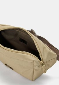 Borsa in tessuto beige con chiusura a zip; interno con rivestimento scuro; include una tracolla marrone e un'etichetta con il marchio.