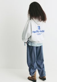 Felpa grigia oversize con testo blu, indossata sopra una maglietta a righe. Pantaloni di jeans larghi con polsini arricciati, abbinati a stivali alla caviglia marroni.