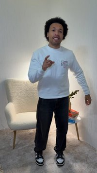 Άσπρο φούτερ με γραφική λεπτομέρεια, σκουρόχρωμο τζιν και μαύρα αθλητικά παπούτσια. Στάση όρθια, ανοιχτόχρωμη καρέκλα και βιβλία στο φόντο.