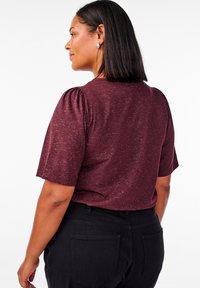 Bordo, teksturirana majica s kratkim rukavima i napuhanim ramenima, uparena s crnim hlačama. Materijal izgleda mekano s blagim sjajem.