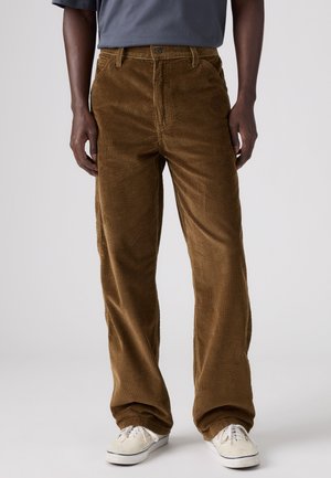 Personne portant un pantalon en velours côtelé marron et des baskets blanches, debout devant un fond clair uni.