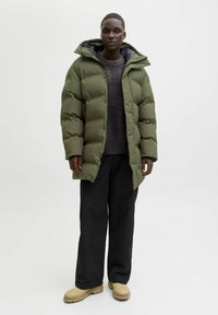 Person som bär en olivgrön vadderad jacka, mörk tröja, svarta vida byxor och beigea stövlar, stående mot en enkel ljus bakgrund.