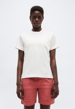 Mujer con camiseta blanca de manga corta y pantalones cortos cargo color coral, de pie contra un fondo claro liso.