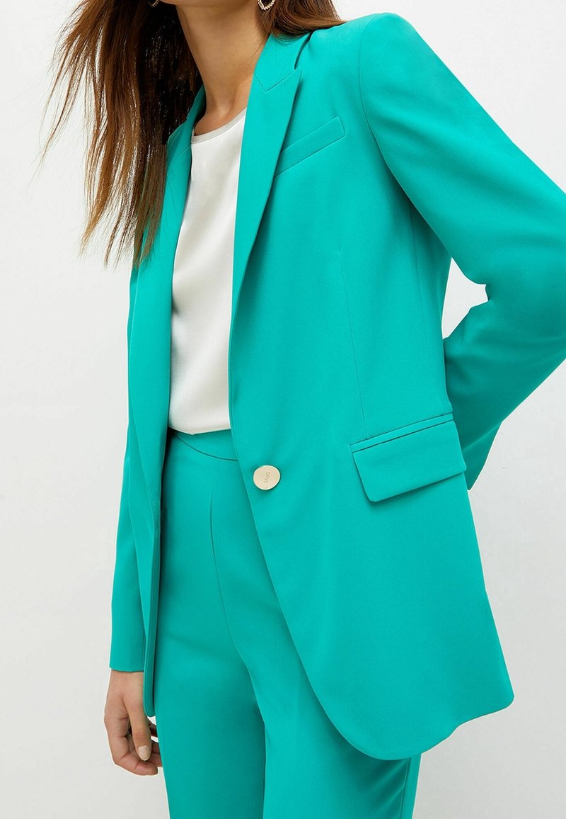 Femme portant un blazer turquoise et un pantalon assorti sur un chemisier blanc, la main droite posée sur la hanche.