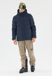Marineblaue Puffjacke mit Reißverschluss, hellbeige Schneehose mit schwarzen Akzenten und übergroßen, verspiegelten Skibrillen. Beanie in hellbraun.