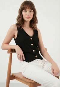 Jeune femme aux cheveux bruns mi-longs, portant un gilet noir à boutons et un pantalon blanc, assise sur une chaise en bois devant un fond uni.