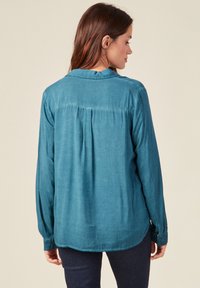 Maglietta turchese a maniche lunghe con una texture morbida, polsini con bottoni, una piega sul retro e un orlo arrotondato. Cuciture dettagliate lungo il colletto.
