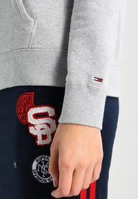 Sudadera gris con puños acanalados, que presenta un pequeño logo en rojo, blanco y azul. Pantalones de chándal azul oscuro con parches rojos y blancos.