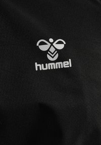 Logo blanc d'abeille stylisée au-dessus du texte en minuscules « hummel » sur tissu noir texturé, indiquant l'emblème d'une marque de vêtements de sport.