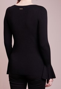 Svart ribbad tröja med långa ärmar och utsvängda muddar. Har en bred halsringning och en liten metallaccent på baksidan.