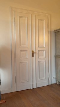 Kahekordse paneeliga valge puidust uks, vertikaalsete soontega, kuldse käepideme ja hingedega. Paigutatud heledale seinale, raamitud puitpõrandaga.