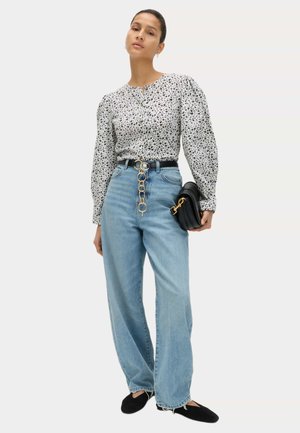 Donna che indossa una camicetta bianca a fiori con maniche a sbuffo, jeans azzurri a vita alta, ballerine nere e tiene in mano una borsa nera con dettagli dorati.