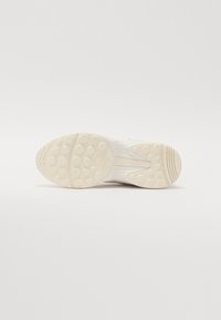 Adidași albi cu o talpă texturată ce prezintă modele circulare și orizontale. Logo-ul fabricantului este vizibil în zona călcâiului.