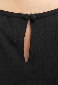 Tissu noir à surface texturée, avec une petite ouverture en forme de serrure au dos et une fermeture par bouton juste au-dessus.