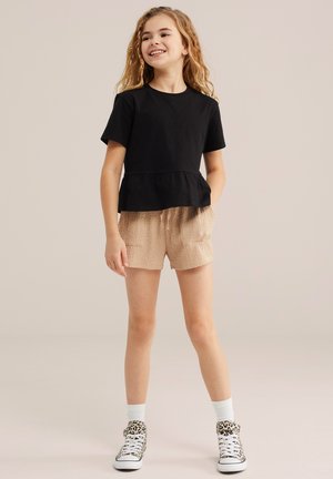 Niña joven con cabello largo y rizado, vestida con una camiseta negra de manga corta, pantalones cortos beige con textura, calcetas blancas y zapatillas con estampado de leopardo, sonriendo y de pie.