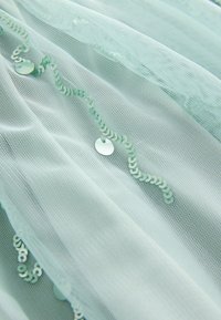 Šviesiai mėtų spalvos tiulis su tekstūruotu raštu, kuriame visame paviršiuje eina banguota blizgių, apvalių pailgų blizgučių linija, panašios spalvos.