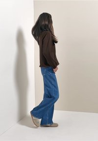 Maglietta marrone a maniche lunghe abbinata a jeans ampi blu, su uno sfondo neutro. L'outfit include scarpe da ginnastica beige.