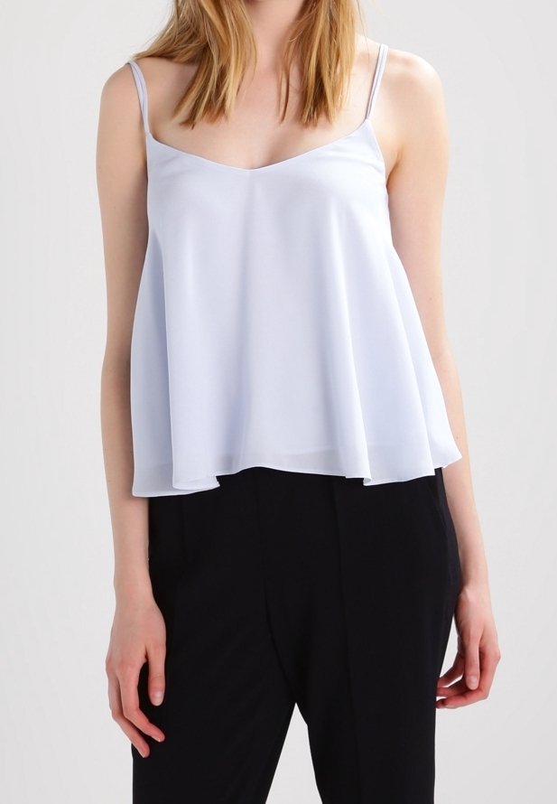 Femme portant un débardeur ample bleu clair à fines bretelles et un pantalon noir sur un fond uni.