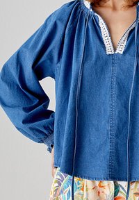 Blusa di jeans con scollatura a V, bordo in pizzo bianco, maniche lunghe elasticizzate e dettagli con cordoncino; vestibilità rilassata e colore blu medio.