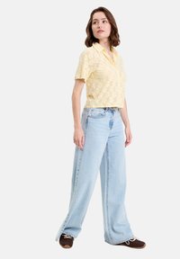 Chemise à manches courtes, jaune clair, avec une texture florale, associée à un jean bleu clair, décontracté et ample, et des baskets marron. Couleurs unies avec une coupe décontractée.