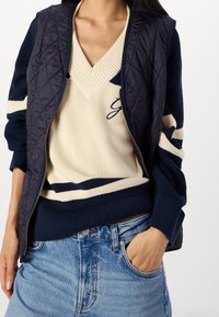 Prošívaná námořnicky modrá vesta p�řes krémový svetr s výstřihem do V a modro-krémovými pruhy, doplněný světle modrými džínami.