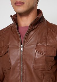 Een bruin leren jack met een ritssluiting, hoge kraag en twee borstzakken. Het materiaal lijkt glad met een lichte glans.