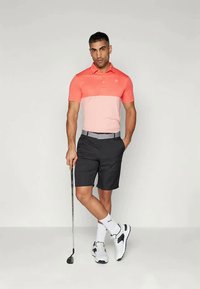 Ανδρική στολή γκολφ με πόλο χρωματικού μπλοκ σε ροζ και κοραλλί, μαύρα σορτς και λευκά αθλητικά παπούτσια με γκρι λεπτομέρειες. Ο άνδρας κρατάει ένα γκολφ. 