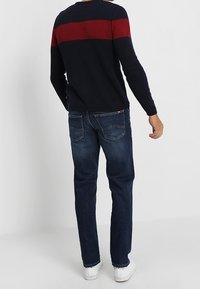 Granatowy sweter dzianinowy z czerwoną poziomą paską, zestawiony z ciemnymi dżinsami. Białe sneakersy. Tekstura ukazuje ściągacze oraz klasyczne detale dżinsowe.