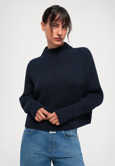 Marineblauwe gebreide trui met hoge kraag en ribbelstructuur, voorzien van lange mouwen en een cropped ontwerp, gecombineerd met lichtblauwe jeans.