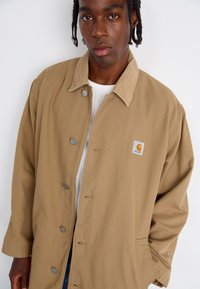Beige button-up jakke lavet af slidstærkt stof, med en kordfløjlskrave, trykknaplukninger og et firkantet logo-patch på brystet.